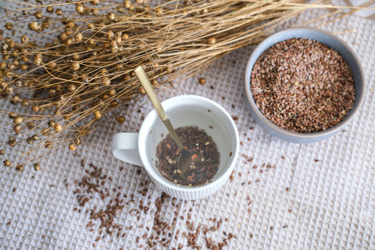 A cup of water and golden teaspoon of flax seeds in a bowl and dried flax flowers on a cotton napkin background. Organic food, veganism, vitamins, health.