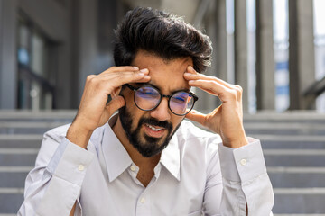 A close-up photo of a young Indian male office worker sitting outside a building on the steps and holding his head in his hands, feeling severe pain and pressure.