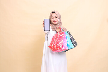a young Asian Muslim girl wearing a hijab smiles gracefully displays a cellphone screen and carries a paper bag. for advertising, technology, Eid and Ramadan