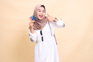 a beautiful Asian Muslim woman wearing a hijab smiles cheerfully with a thumbs up gesture okay and brings a debit credit card for online offline payments. used for advertising, technology and Ramadan
