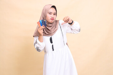 Fototapeta premium a beautiful Asian Muslim woman in a hijab frowns with a thumbs up gesture and carries a debit credit card for offline online payments. used for advertising, technology, Eid and Ramadan