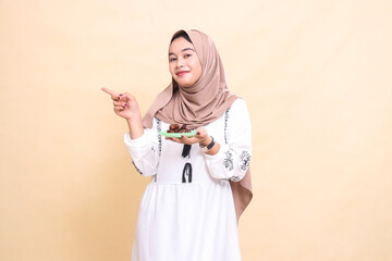 a beautiful Asian Muslim woman wearing a hijab smiles, pointing to the right and carrying dates for the sahur menu and breaking the fast. used for advertising, health, Eid and Ramadan