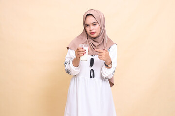 A beautiful indonesia Muslim woman wearing a hijab with an angry expression carries and points to a glass of white milk for eating and breaking the fast. used for advertising, health, Eid and Ramadan