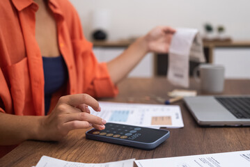 Expense Management: Seated at a desk with a laptop, a woman uses her smartphone calculator to...
