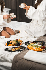 Obraz na płótnie Canvas Young women in bathrobes have breakfast in bed, drink coffee and fool around. Breakfast cheesecakes and fruits with coffee. Hotel holiday concept
