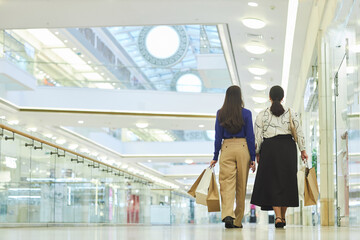 Back view of two women walking away from camera in shopping mall and holding bags enjoying weekend...