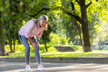 Senior gray-haired woman walks and does sports in the park, stands bent over in pain, holds hands...