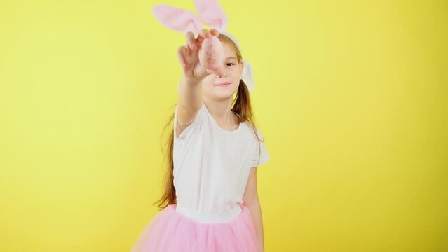 Portrait child in bunny ears, looking at camera. Cute little girl pointing on colored egg, posing isolated over yellow color background.