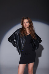 Stylish beautiful fashionable woman in fashion clothes with a dress and leather jacket on a dark background in the studio