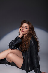 Beautiful fashion stylish woman model with hairstyle in a fashionable dress with a leather jacket sits on the floor in the studio