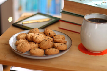 Cup of tea or coffee, pile of books, plate of cookies, reading glasses, e-reader and pen on the table. Colorful rainbow bookshelf in the background. Selective focus.
