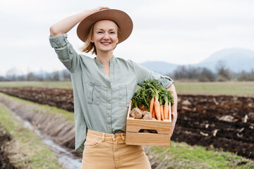 Female farmer holding wooden box full of fresh raw vegetables in agricultural field area. Woman standing proud with freshly bunch harvest outdoors. - 768746487