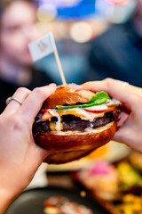 person’s hands hold a juicy burger with melted cheese, lettuce, and sauce. The glossy bun is...