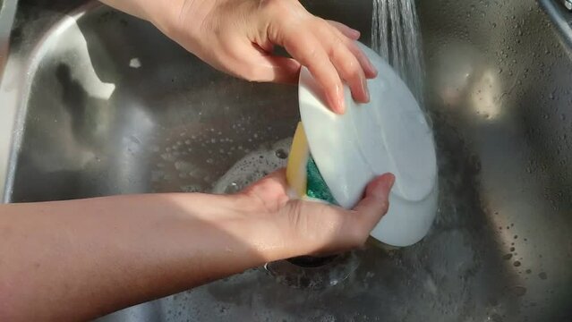 Unas manos de  mujer lavando un plato de color blanco  con una esponja amarilla con jab&oacute;n liquido y agua  en un fregadero