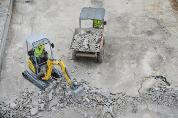 Workers operating a mini excavator machine removes the old concrete with the shovel and deposits it...