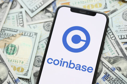 KYIV, UKRAINE - MARCH 15, 2024 Coinbase logo on iPhone display screen with many hundred dollar bills. Cryptocurrency exchange portal