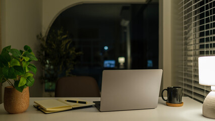 Laptop computer, stationery, coffee cup and light from table lamp in dark office at night.
