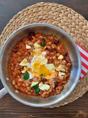 shakshuka, white beans in tomatoes, fried eggs with beans and tomatoes, wicker placemat