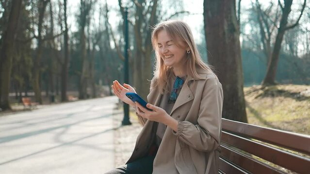 Happy smiling young blonde woman sitting in nature green park outdoors using mobile phone. Winning woman playing game, paying bills, good shopping. High quality FullHD footage