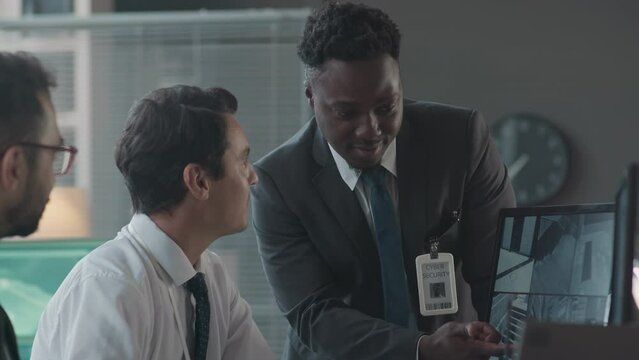 Tilt up shot of Black male cybersecurity officer and his diverse male colleagues discussing multiple CCTV cameras on computer monitor while co-working in modern office