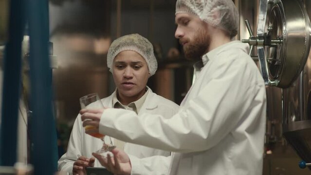 Medium shot of two ethnically diverse brewery workers in lab coats smelling and tasting freshly brewed beer poured in glass from fermenter tank