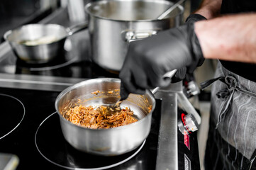 In this professional kitchen, a chef in black gloves is skillfully stirring ingredients in a stainless steel pan. The focus is on their hands, emphasizing the cooking action
