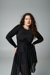beautiful plus size young girl in black outfit  posing with hands on hips on grey background