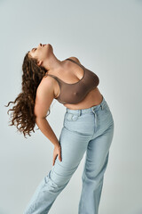 alluring plus size woman in brown bra and blue jeans with curly hair putting head to behind - obrazy, fototapety, plakaty