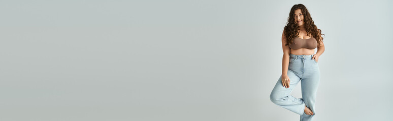 banner of cheerful plus size woman in brown bra and blue jeans with curly hair posing with bent leg