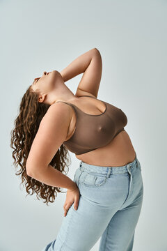 plus size young woman in brown bra and blue jeans with curly hair putting head to behind with hand