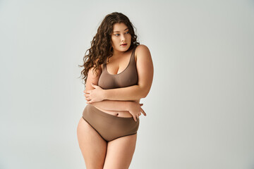 beautiful plus size young girl in brown lingerie with curly hair hugging herself and looking to side - obrazy, fototapety, plakaty