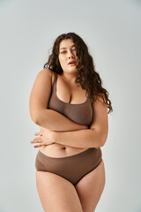 alluring plus size young woman in underwear with curly brown hair hugging herself on grey background - 768801015