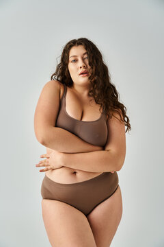 Fototapeta alluring plus size young woman in underwear with curly brown hair hugging herself on grey background