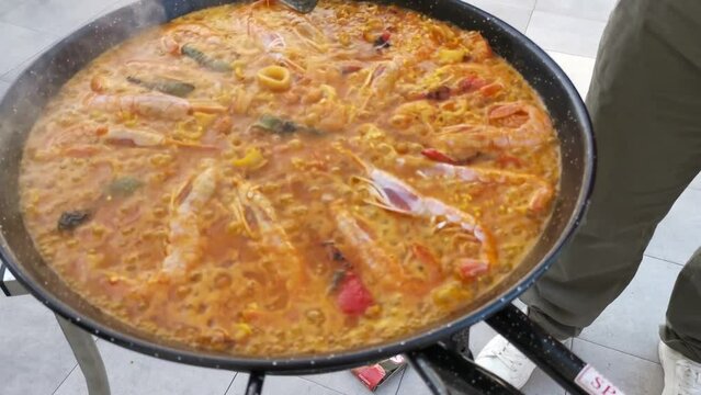 Man holding a ladle flipping a big Paella Valenciana dish inside coocking bowl