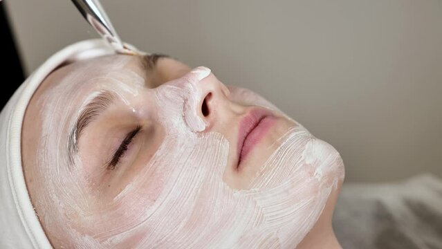 professional cosmetologist applies a white mask on the face of a female client with a brush, close-up