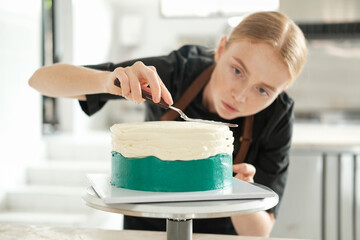 Portrait of a young red haired female pastry chef who looks at the cake with concentration and distributes the cream in an even layer over its surface. The process of preparing in a production kitchen