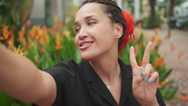 Young caucasian pretty woman with red dreadlocks showing V sign, making selfie with happy smile on background exotic plants