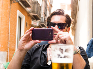 Spanish Caucasian man tourist with sunglasses laughs and takes a photo with his cell phone on the terrace of a Spanish bar - 768851605