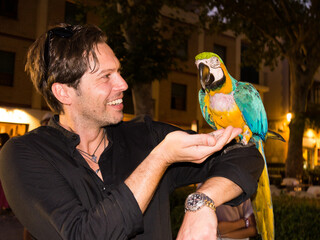 Laughing caucasian handsome man in black shirt holds and looks at a parrot and feeds it on the street at night - 768852281