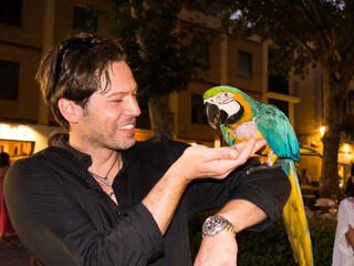 Laughing caucasian handsome man in black shirt holds a parrot and feeds it looking at his hand on the street at night - 768852427