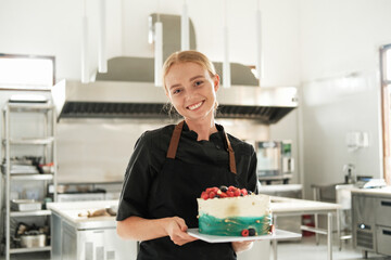 Portrait of a young charming red haired female pastry chef who stands in an apron against the backdrop of the production workshop. In front of her on the table is a cake decorated with fresh berries.