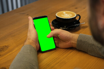 Young man sitting at cafe holding smartphone green mock-up screen in hand. Male person using chroma...