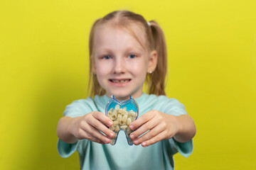 A beautiful seven year old girl holds an artificial tooth against a yellow background. The concept of pediatric orthodontics, malocclusion and early tooth decay in children, close-up