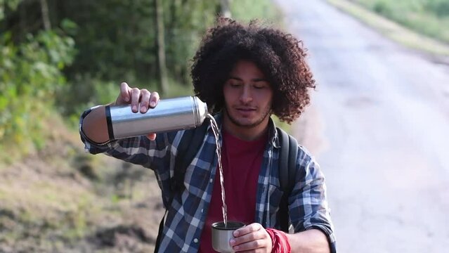 a young black-haired Arab guy pours boiling water from a thermos into a mug against the backdrop of forest, hot tea on a walk. High quality FullHD footage