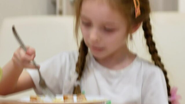 A little girl of 6 years old eats pasta at the table at home in a bright interior, with disheveled pigtails. The noodles are hanging out of my mouth.
