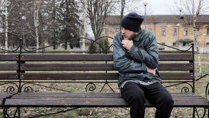 Bearded homeless man sits on bench in city park, tramp is trying to keep warm from cold outside....