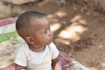 single african toddler , with a fly on his face, sitting outdoors on a blanket in front of the...