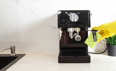 A girl s hand in a yellow glove washes a black coffee machine with a sponge with foam and cleaning agent in the kitchen against a background of a white wall with porcelain tiles. Copy space for text. 
