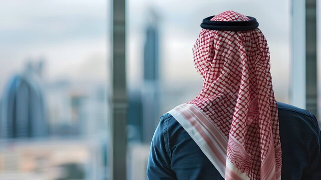 Arab man wearing Saudi bisht, looking at office window looking at city view, back view, blurred background.