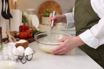 Woman making dough with whisk in bowl at light table, closeup